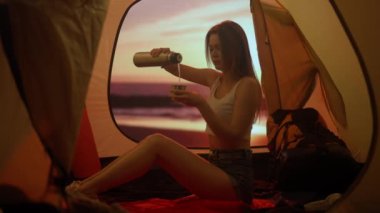 Kamp ve macera konsepti. Kamp alanında gezen, yürüyüş yapan, doğayı keşfeden biri. Genç bir kadın termostan çay dolduruyor. Gün batımında sahilde çadırın içinde oturuyor..