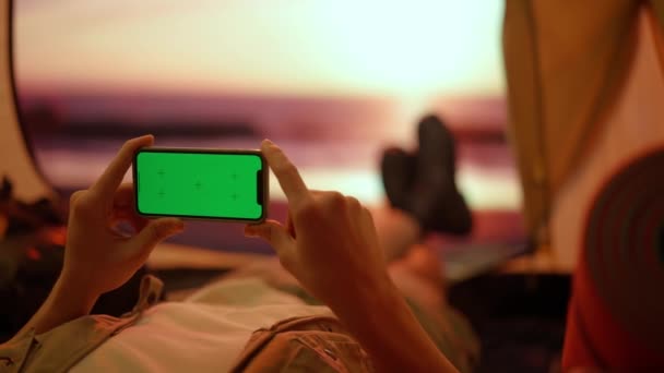 露营和探险的概念 人在营地旅行和远足 探索自然 年轻男子躺在沙滩上的帐篷里 带着智能手机色钥匙绿色屏风 外出度假 — 图库视频影像