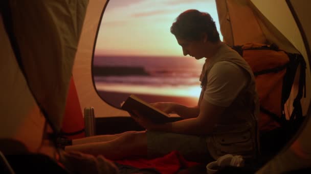 キャンプと冒険のコンセプト キャンプ場の旅行やハイキング 自然を探検する人 ビーチ 屋外での日没時にテントの中の本を読んでいる若い男 — ストック動画