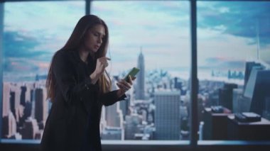 Şirket reklam konsepti. Ofiste başarılı bir iş kadını. Akıllı telefonlu kadın koruma fonu yöneticisi, şehir manzaralı pencerenin önünde kazanan jesti gösteriyor.