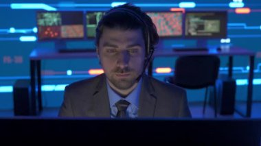 Proje yöneticisi bilgisayar bilimi konsepti. Kontrol odasında teknik destek. İnsan bilişim uzmanı ofiste kulaklıkla konuşan bir bilgisayar üzerinde çalışıyor. Büyük dijital ekranda altyapı verileri görülüyor..