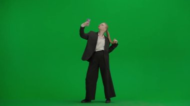 Modern iş kadını yaratıcı reklam konsepti. Krom anahtar yeşil ekranda takım elbiseli kadın portresi. Resmi kıyafetli sarışın iş kadını elinde akıllı telefonuyla selfie çekiyor..