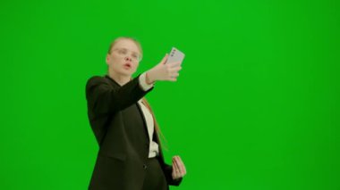 Modern iş kadını yaratıcı reklam konsepti. Krom anahtar yeşil ekranda takım elbiseli kadın portresi. Resmi kıyafetli sarışın iş kadını elinde akıllı telefonuyla selfie çekiyor..