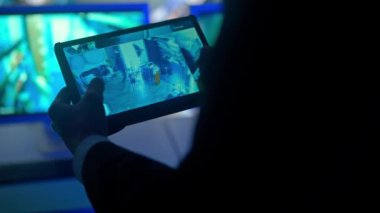 Gözetleme ve güvenlik konsepti. Güvenlik görevlisi, izleme merkezinde tablet üzerinde çalışıyor. Görüntüyü yakınlaştırıyor. Bilgisayarlar ve büyük dijital ekran kamera görüntüleri.