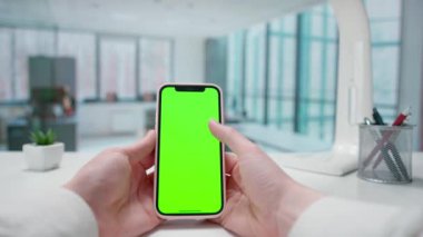 Mans elleri akıllı telefon yeşil ekran kroma anahtarıyla etkileşimde. Pencereleri olan parlak bir ofis. Metin veya resim için şablon yeri, promosyon içeriği. Reklam alanı, çalışma alanı maketi.