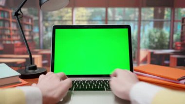 Laptop klavyesinde yeşil ekran krom anahtarı olan bir adam. Pencereli sıcak kütüphane arkaplanı. Metin veya resim için şablon yeri, promosyon içeriği. Reklam alanı çalışma alanı modeli