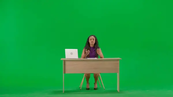 Tv news and live broadcasting concept. Female in dress sit at the desk isolated on chroma key green screen background. Full shot african american woman tv news host sitting talking looking at camera.