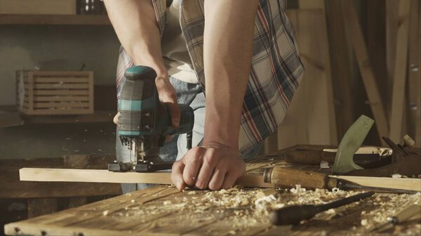 Carpentry and handicraft advertisement concept. Male woodworker working in garage. Man professional carpenter working with wooden materials in workshop sawing wood with jigsaw.