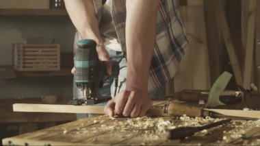 Marangozluk ve el işi reklam konsepti. Garajda çalışan erkek marangoz. Profesyonel marangoz atölyede ahşap malzemelerle çalışıyor. Yapboz ile odun kesiyor..