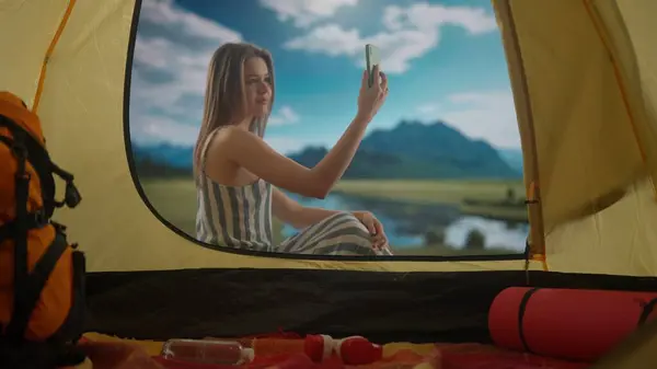 キャンプと冒険のコンセプト 湖の近くでリラックスしたキャンプ場で休む女性 テントの外に座っている若い女性は 山岳湖の海岸で昼光を浴び スマートフォンでビデオ通話で話す — ストック写真
