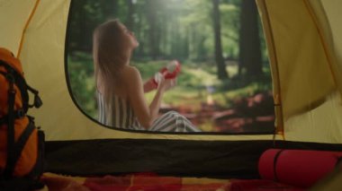 Kamp ve macera konsepti. Kadın kamp alanında dinleniyor, vahşi doğada dinleniyor. Ormanda gün ışığında çadırın dışında oturan, kulaklıkla müzik dinleyen, dışarıda tatil yapan genç bir kadın..