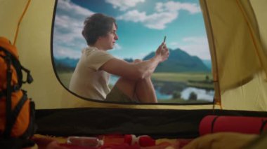 Kamp ve macera konsepti. Kamp alanında dinlenen bir erkek göl kenarında dinleniyor. Genç adam gün ışığında dağların kıyısında çadırın dışında oturuyor, akıllı telefondan video ile konuşuyor.,