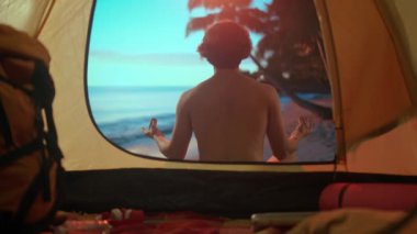 Kamp ve macera konsepti. Erkek, kamp alanında dinleniyor, okyanus kenarında dinleniyor. Genç adam gün batımında çadırın dışında oturuyor plajda meditasyon yapıyor, dışarıda tatil yapıyor..