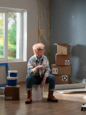 Yaşlı bir kadın, evin tadilatı sırasında etrafı kutularla çevrili bir boya kovasıyla düşünceli bir mola veriyor..