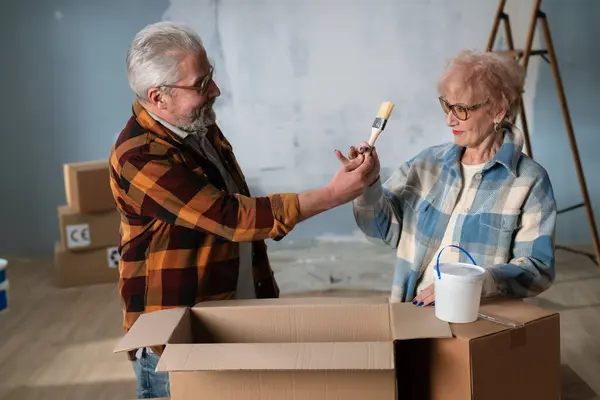 一对年长的夫妇喜欢在家居翻新过程中打开包装盒 分享快乐的时光 一位老人拿出画笔 很高兴地把它拿给他的妻子看 — 图库照片