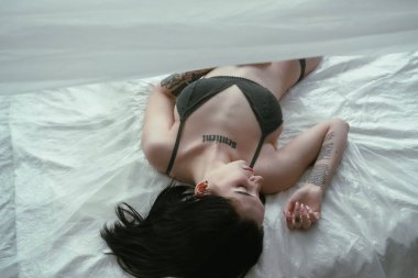 Dövmeleri olan bir kadın beyaz bir yatakta uzanmış, huzur veren bir güzelliği taşıyor.