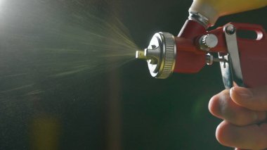 Atölyeye sarı boya sıkan kırmızı boya tabancasının detaylı görüntüsü. İnce boya parçacıkları sprey silahtan püskürtülür ve farklı yönlere uçar..
