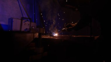 Metal bir ürün üzerinde çalışan bir kaynakçının yöntemi. Kaynakçı demir yüzeyde kaynak makinesi, duman, kıvılcımlar ve yanıp sönen ışıklar kullanıyor. Mavi ışıklı karanlık bir atmosfer..