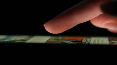 Düşük ışıkta bir akıllı telefon ekranında sosyal medyada kayan bir parmağın yakın çekimi, aygıtların dokunmatik ekranla etkileşimini vurguluyor.