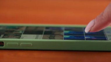 Tahta bir masadaki akıllı telefon ekranında, bir galeri yayını boyunca ilerleyen bir eli kapatın, cihaz ile etkileşime önem verin..