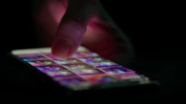 Düşük ışıkta bir akıllı telefon ekranında fotoğraf galerisinde kaydırılan bir parmağın yakın çekimi, aygıtların dokunmatik ekranla etkileşimini vurguluyor.