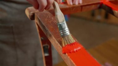 Bir atölyedeki ahşap sandalye yüzeyine kırmızı boya sürmek için fırça kullanan bir elin yakın çekimi, boyama ve ahşap işçiliği sürecini gözler önüne seriyor..