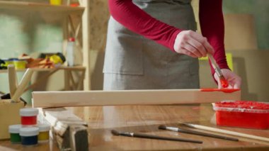 Bir atölyedeki ahşap bir yüzeye kırmızı boya sürmek için fırça kullanan bir elin yakın çekimi boyama ve ahşap işçiliği sürecini gözler önüne seriyor..