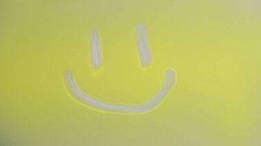 Camdan bir yüzey üzerinde sarı bir parmak ile çizilmiş gülen bir yüz resmi yakın çekim. Basit bir tasarım neşeli ve olumlu bir duygu taşır..