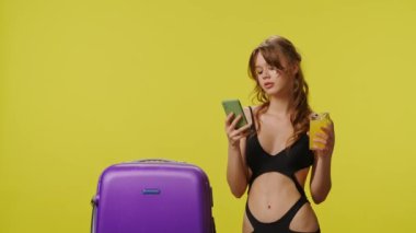 Bavulla poz veren, akıllı telefondan mesaj atan mayo giymiş genç bir kadın. Seyahat, yaz tatili, plaj tatili konsepti. Sarı arka planda siyah bikinili oyuncu genç bayan model..