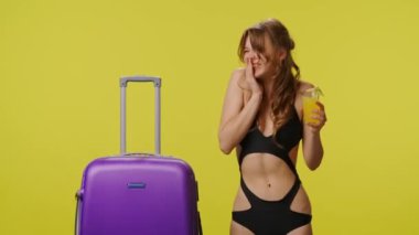 Bavullu ve kokteylli genç bir kadın heyecanlı bir ifade takınıyor. Seyahat, yaz tatili, plaj tatili konsepti. Sarı arka planda bikinili oyuncu bir bayan model.