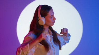 Gündelik giysiler içinde şık genç bir kadın kulaklıkla müzik dinliyor, akıllı telefon kullanıyor ve dans ediyor. Renkli stüdyo ışıkları altında izole edilmiş. Kadın model renkli ışıklarda kameraya poz veriyor