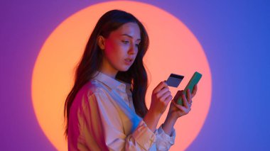 Günlük kıyafetlerle şık genç bir kadın akıllı telefon uygulamasında banka kartı numarası yazıyor, online ödeme. Renkli bir stüdyo geçmişiyle izole edilmiş. Renkli ışıklarda kameraya poz veren kadın model..