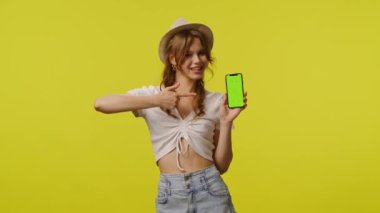 Gündelik giyinen genç bir kadın başparmağında akıllı telefon kroma anahtar yeşil ekran tutuyor. Seyahat, yaz tatili, turizm eğlence konsepti. Sarı arka planda izole edilmiş kameraya poz veren oyuncu kadın model.