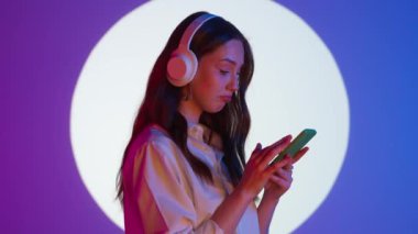 Gündelik giysiler içinde şık genç bir kadın kulaklıkla müzik dinliyor ve akıllı telefondan internette geziniyor. Renkli stüdyo ışıkları altında izole edilmiş. Kadın model renkli ışıklarda kameraya poz veriyor