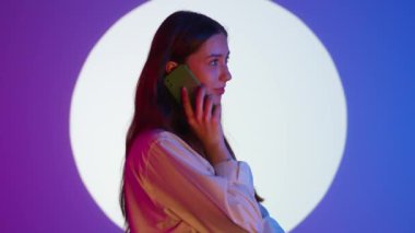 Gündelik giysiler içinde şık genç bir kadın akıllı telefondan arıyor. Renkli stüdyo ışıkları altında izole edilmiş. Renkli ışıklarda kameraya poz veren kadın model..
