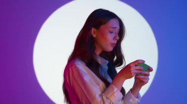 Gündelik giysiler içinde şık genç bir kadın akıllı telefondan video oyunu oynuyor ve mutlu bir yüz kazanıyor. Renkli stüdyo ışıkları altında izole edilmiş. Renkli ışıklarda kameraya poz veren kadın model..