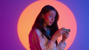 Gündelik giysiler içinde şık genç bir kadın akıllı telefondan video oyunu oynuyor ve mutlu bir yüz kazanıyor. Renkli stüdyo ışıkları altında izole edilmiş. Renkli ışıklarda kameraya poz veren kadın model..