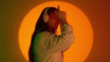 Gündelik giysiler içinde şık genç bir kadın kulaklıkla müzik dinliyor ve dans ediyor. Renkli stüdyo arka planında sarı spot ışıklarıyla izole edilmiş. Renkli ışıklarda kameraya poz veren kadın model..