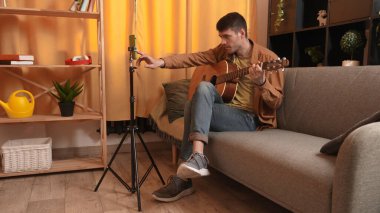 Evde gitar çalan genç bir müzisyen. Yetişkin gitarist odada oturmuş akustik gitar çalıyor, sosyal medya için akıllı telefondan video kaydediyor..