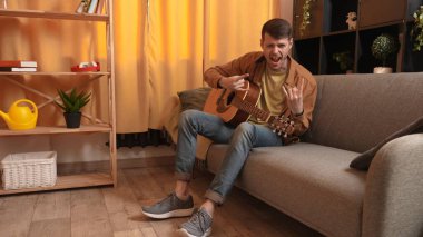 Evinde gitar çalan genç bir müzisyen. Yetişkin bir erkek gitarist bir odada oturur ve bir kaya hareketi gösterir.
