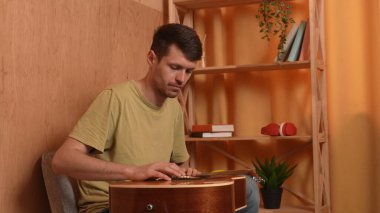 Evde gitar çalan genç bir müzisyen. Yetişkin bir gitarist odada oturup akustik gitar çalıyor. Tahta gövdeye dokunuyor, telli akorları parmaklıyor. İşlemi seviyor..