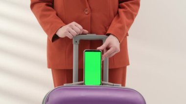 Turuncu elbiseli bir kadın mor bir bavul tutacağı tutuyor ve yeşil ekran akıllı telefonu işaret ediyor.