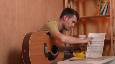 Evde gitar çalan genç bir müzisyen. Yetişkin bir gitarist odada oturmuş notalarla akustik gitar çalıyor ve parmaklama melodisini kontrol ediyor. Hobi ve boş zaman kavramı.