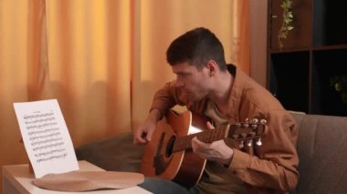 Evde gitar çalan genç bir müzisyen. Yetişkin bir gitarist odada oturmuş notalarla akustik gitar çalıyor ve parmaklama melodisini kontrol ediyor. Hobi ve boş zaman kavramı.