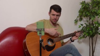 Evde gitar çalan genç bir müzisyen. Yetişkin gitarist odada oturmuş akustik gitar çalıyor, sosyal medya için akıllı telefondan video kaydediyor..