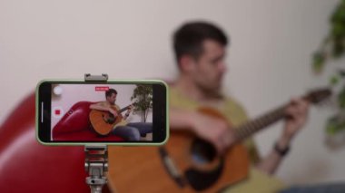 Evde gitar çalan genç bir müzisyen. Yetişkin bir gitarist kanepede oturmuş akustik gitar çalıyor, akıllı telefon kullanarak kendini videoya kaydediyor. Hobi ve boş zaman kavramı.