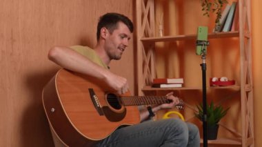 Evde gitar çalan genç bir müzisyen. Yetişkin bir gitarist odada oturmuş akustik gitar çalıyor sosyal medya için akıllı telefondan video kaydediyor. Yavaş çekim.