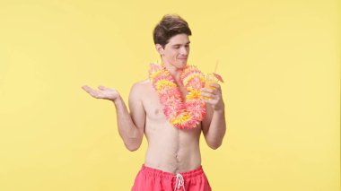 Kokteyl içen genç bir adam. Tadı ve tazeliği onu çok mutlu ediyor. Pembe şortlu çıplak bir adam ve sarı arka planda Hawaii lei pozu. Seyahat, yaz tatili.