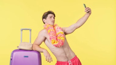 Genç adam akıllı telefonuyla poz veriyor ve selfie çekiyor. Pembe şortlu tişörtsüz bir adam ve sarı arka planda poz veren Hawaii zambağı. Seyahat, yaz tatili.