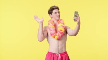 Genç adam akıllı telefon kullanarak görüntülü konuşma yapıyor. Pembe şortlu çıplak gövdeli erkek model ve sarı arka planda Hawaii zambağı pozu. Seyahat, yaz tatili, plaj eğlencesi konsepti.